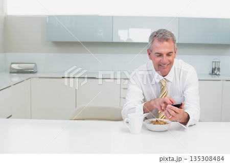 Senior man in business attire holding smartphone while eating cereal at modern kitchen, copy space Senior man in business attire holding smartphone while eating cereal at modern kitchen, copy space 135308484