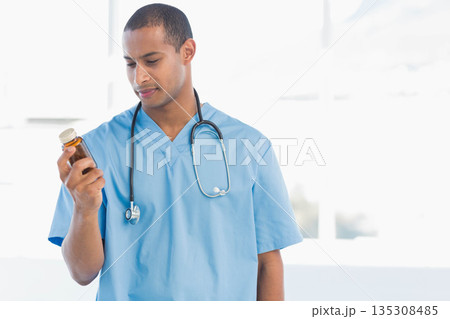 African American male doctor in light blue scrubs and stethoscope examining pill bottle in clinic 135308485