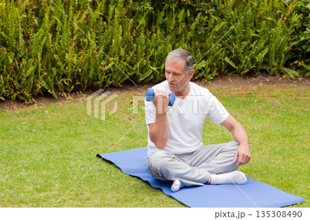 Senior man performing bicep curl on blue exercise mat on grass lawn with blue dumbbell Senior man performing bicep curl on blue exercise mat on grass lawn with blue dumbbell 135308490