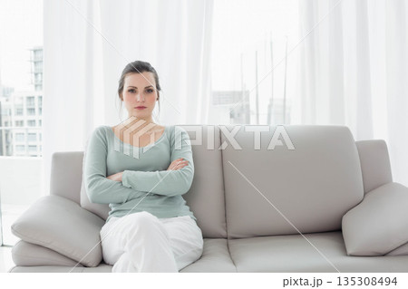 Woman in her twenties sitting on light gray sofa at home folding arms beside sheer curtains 135308494