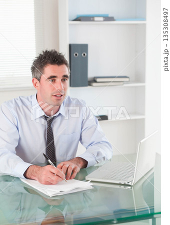 Man in business attire writing on papers at glass-topped desk beside open laptop and binders 135308497