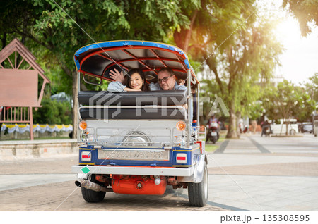 Old white man and asian woman waving a hand while riding in tuk tuk three wheel taxi touring a city. Old white man and asian woman waving a hand while riding in tuk tuk three wheel taxi touring a city. 135308595