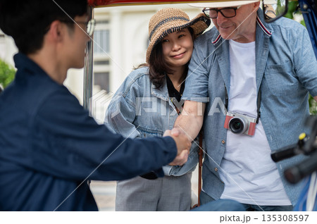 Old white man handshaking a driver aside wife after riding in a tuk tuk or three wheeled taxi touring a city. 135308597