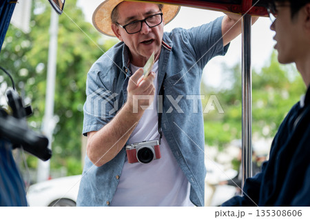 Old white man paying fare to asian driver after riding in tuk tuk or three wheel taxi touring a city Old white man paying fare to asian driver after riding in tuk tuk or three wheel taxi touring a city 135308606