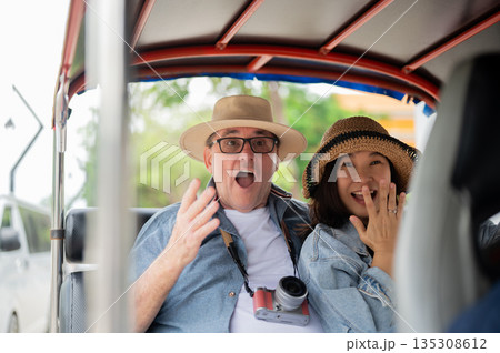 Old white man and asian woman looking surprised as riding in tuk tuk three wheel taxi touring a city 135308612