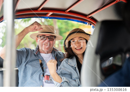 Happy couple old white man and asian woman sitting riding in tuk tuk three wheel taxi touring a city Happy couple old white man and asian woman sitting riding in tuk tuk three wheel taxi touring a city 135308616