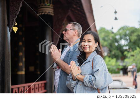 Old white man and smiling asian woman joining palms praying worshipping at ancient buddhist temple. 135308629