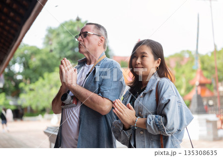Old white man with asian woman joining palms and praying or worshipping at ancient buddhist temple. 135308635
