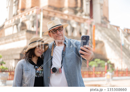 Old white man holding phone taking photos with asian woman standing aside ancient pagoda in a temple 135308660