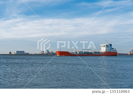 Orange tanker ship sailing on sunny day with blue sky and cityscape on the horizon. 135310063