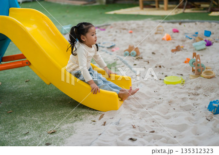 child girl playing and sitting at the bottom of yellow slide in a sandy playground. child girl playing and sitting at the bottom of yellow slide in a sandy playground. 135312323