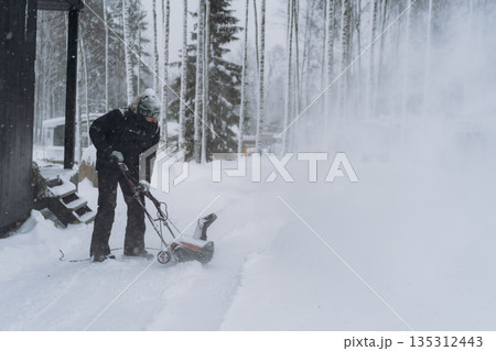 man clearing snow near wooden house in countryside using electric snow blower 135312443