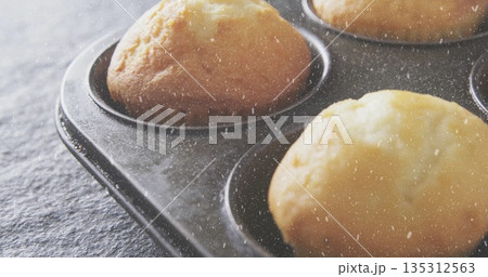 Displaying close-up metal muffin tin holding golden-brown muffins on dark kitchen countertop 135312563