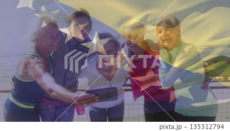 Smiling five women in athletic wear leaning on oceanfront railing, with smartphone and flag overlay 135312794