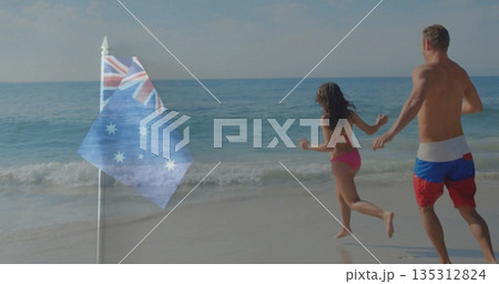 Running couple in trunks and pink bikini splashing along beach shoreline, with Australian flagpole 135312824