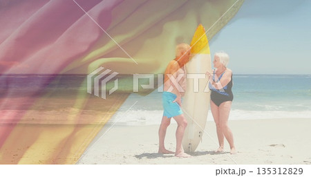 Standing senior couple wearing swim trunks and one-piece facing ocean, with yellow-tipped surfboard Standing senior couple wearing swim trunks and one-piece facing ocean, with yellow-tipped surfboard 135312829
