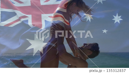 Man lifting partner overhead in swimwear on beach at shoreline, with fading Australian flag overlay 135312837