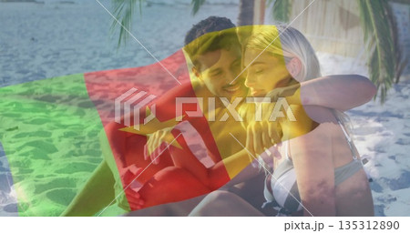 Sitting couple in swimwear embracing each other on sandy beach near calm sea, with flag overlay 135312890