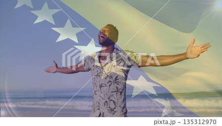 Man wearing tropical shirt raising arms and tilting head back on sandy beach, flag motif overlay 135312970