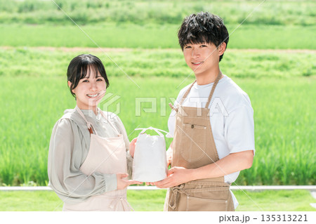 田んぼの前で米袋を持つ米農家の若い日本人夫婦 田んぼの前で米袋を持つ米農家の若い日本人夫婦 135313221