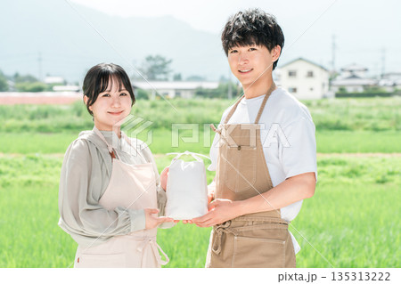田んぼの前で米袋を持つ米農家の若い日本人夫婦 135313222