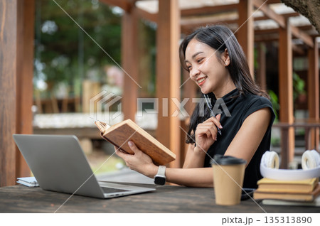 Smiling asian woman student holding pen and book as looking at laptop while sitting at wooden table in outdoor cafe Smiling asian woman student holding pen and book as looking at laptop while sitting at wooden table in outdoor cafe 135313890