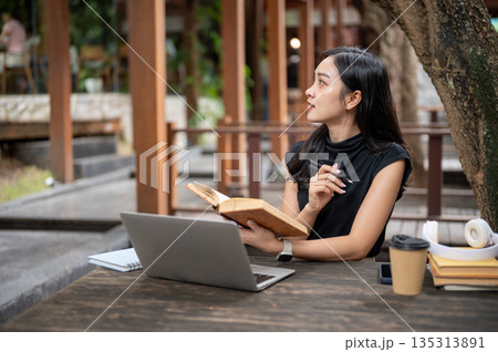Asian woman student holding pen and book as she looking way from laptop while sitting at wooden table in outdoor cafe 135313891
