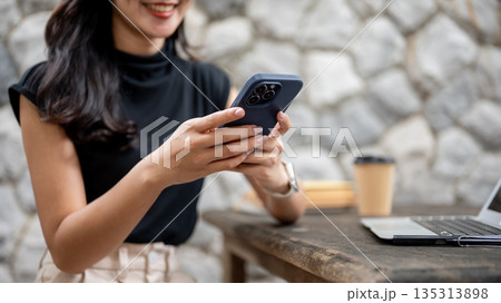 Asian woman looking at phone chatting or playing a game aside laptop while sitting at wooden table in stone wall cafe 135313898