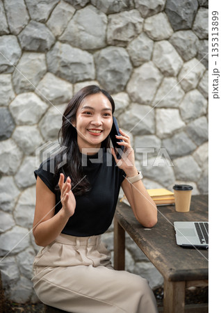 Asian woman smiling during a phone call aside laptop and coffee cup while sitting at wooden table in stone wall cafe 135313899