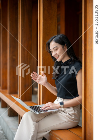 Asian woman looking and raising hand in front of a laptop while sitting on wooden bench in cafe's corridor or hallway 135313905