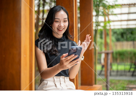 Asian woman looking at phone and raising a hand explaining while sits in cafe's wooden corridor beside bamboo garden Asian woman looking at phone and raising a hand explaining while sits in cafe's wooden corridor beside bamboo garden 135313918
