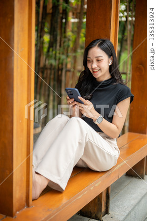Asian woman holding phone and sitting leaning against wooden pillar in cafe's corridor beside bamboo garden 135313924