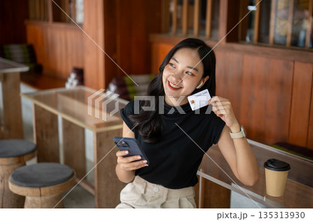 Asian woman holding credit card and looking away from phone while sitting at wooden table in cafe or restaurant 135313930