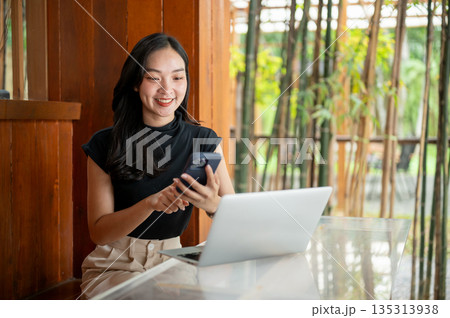 Asian woman holding and looking at phone in front of a laptop while sitting in a cafe or restaurant. Asian woman holding and looking at phone in front of a laptop while sitting in a cafe or restaurant. 135313938