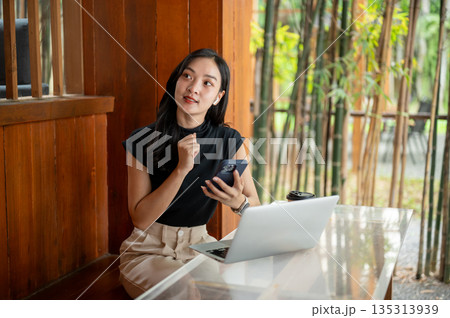 Asian woman holding and looking away from phone while sits in front of a laptop in cafe or restaurant Asian woman holding and looking away from phone while sits in front of a laptop in cafe or restaurant 135313939