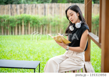 Asian woman holding pen and book reading or writing on wooden bench in a cafe or garden pavilion Asian woman holding pen and book reading or writing on wooden bench in a cafe or garden pavilion 135313950
