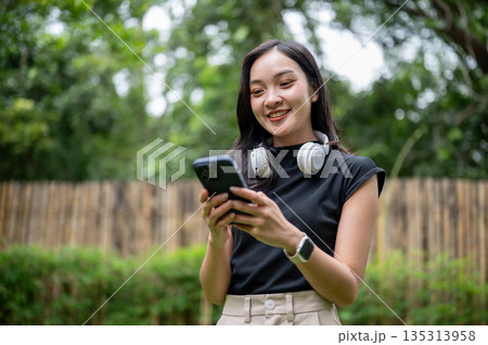 Smiling asian woman holding and looking at phone while standing in her home's garden or outdoor cafe 135313958