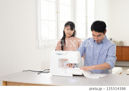 小学生の娘の隣でミシンがけをする父親 135314386