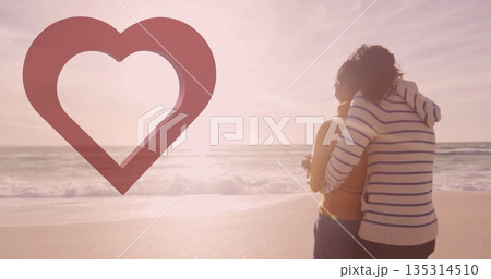 Embracing couple wearing sweater and orange tank on beach, with waves, heart graphic, copy space 135314510