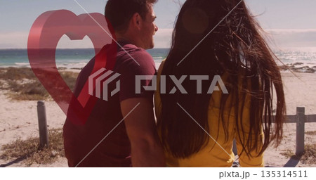 Couple in tshirt, sweatshirt gazing at sea from beach railing posts, with translucent heart graphic 135314511