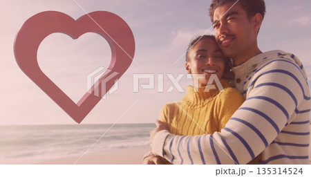 Embracing couple wearing mustard navy-striped sweaters on sandy beach, with ocean waves, heart icon 135314524