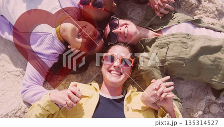 Smiling group of four women lying in circle on sandy beach, with bright sunglasses, summer clothing 135314527