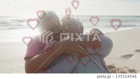 Embracing senior couple sitting on beach edge, displaying wedding rings and heart-shaped graphics Embracing senior couple sitting on beach edge, displaying wedding rings and heart-shaped graphics 135314548