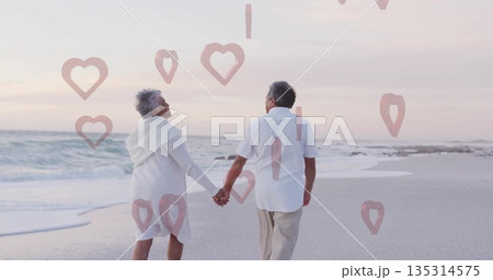 Walking senior couple holding hands along beach at dusk on wet sand, with heart-shaped graphics 135314575