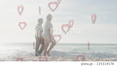 Walking senior couple strolling barefoot at sandy shore with soft surf, digital pink heart overlays Walking senior couple strolling barefoot at sandy shore with soft surf, digital pink heart overlays 135314576