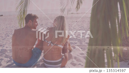 Relaxing adult couple wearing swimwear sitting on beach at sunset, with palm fronds and ocean waves 135314597