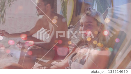 Relaxing couple wearing trunks and bikini top sitting on beach chairs under palms, with surfboard 135314650
