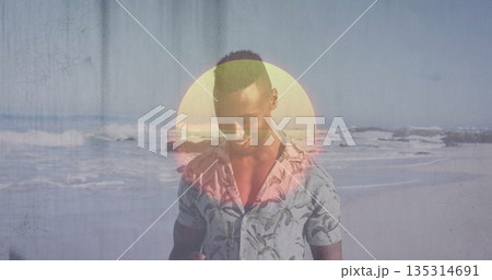 Man in tropical-print shirt holding smartphone on sandy beach shoreline, with pastel sun graphic 135314691