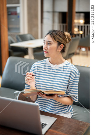 Asian young woman holding pen looking away from book while sits on sofa across laptop at table in cafe. 135315102