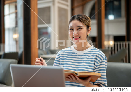 Smiling asian young woman holding pen and book as sit on sofa in front of a laptop on table in cafe. 135315103